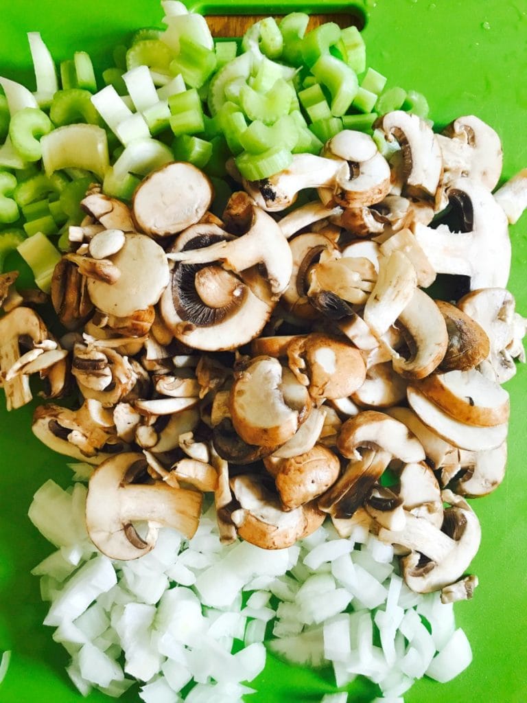 Raw diced celery, mushrooms, and onions on a green cutting board, ready to saute.