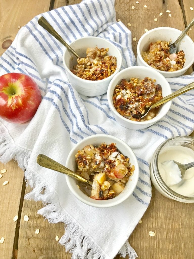 Four white ramekins of Instant Pot apple crisp sitting on a wooden background with coconut cream and an apple sitting nearby 