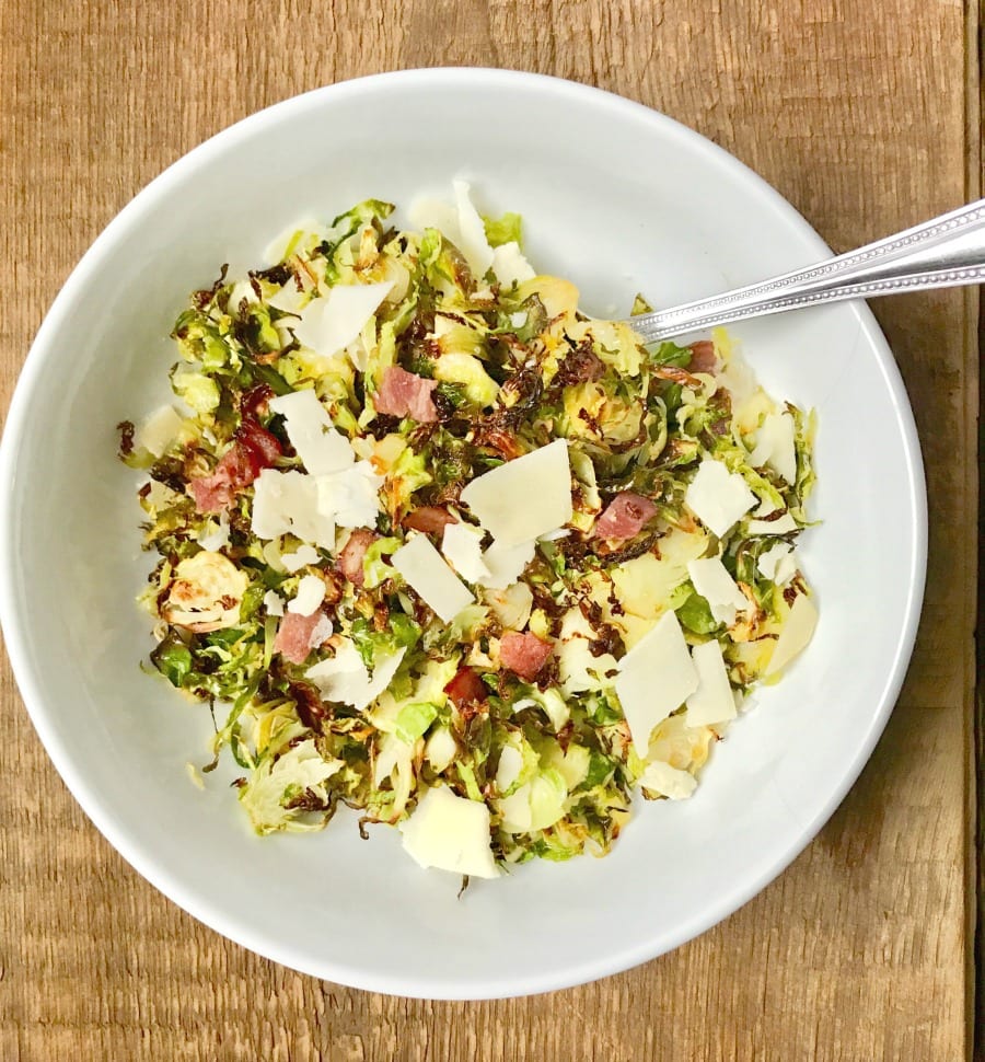 Overhead shot of a large white serving bowl with a silver spoon on a wooden surface. In the bowl is crispy shaved brussels sprouts topped with diced turkey bacon, shaved parmesan cheese, and a balsamic vinegar reduction.