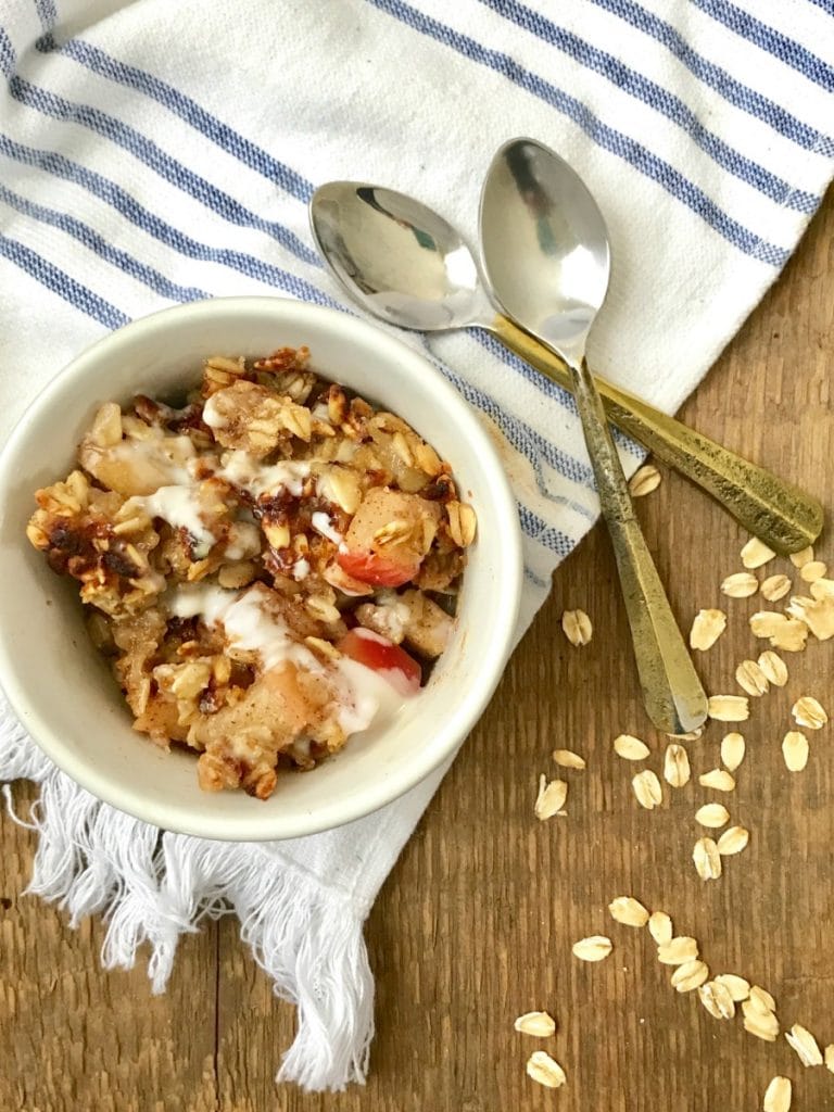 A small ramekin of Instant Pot Apple Crisp sitting on a white and blue striped kitchen towel. Two small spoons sit nearby. 