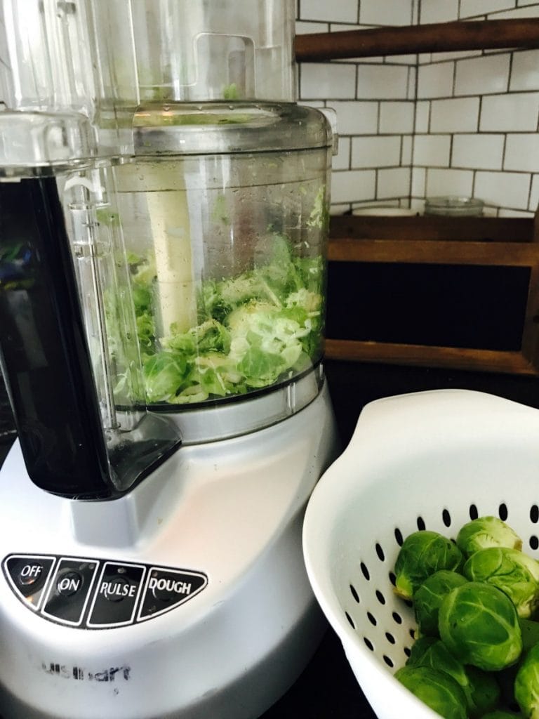 White Cuisinart food processor filled with already shaved brussels sprouts. On the side is a white colander with whole brussels sprouts. In the background there is a white kitchen backsplash and wooden shelf.