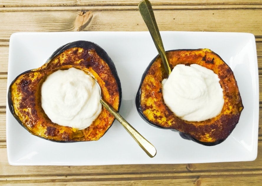 two halves of a roasted acorn squash are sitting on a rectangular plate and stuffed with sweet maple whipped ricotta. There are spoons in each dip, ready to eat!