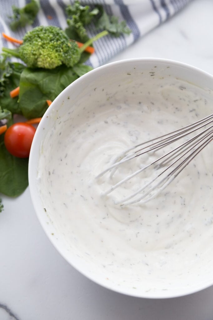 A bowl of homemade ranch dressing sitting next to broccoli and spinach 