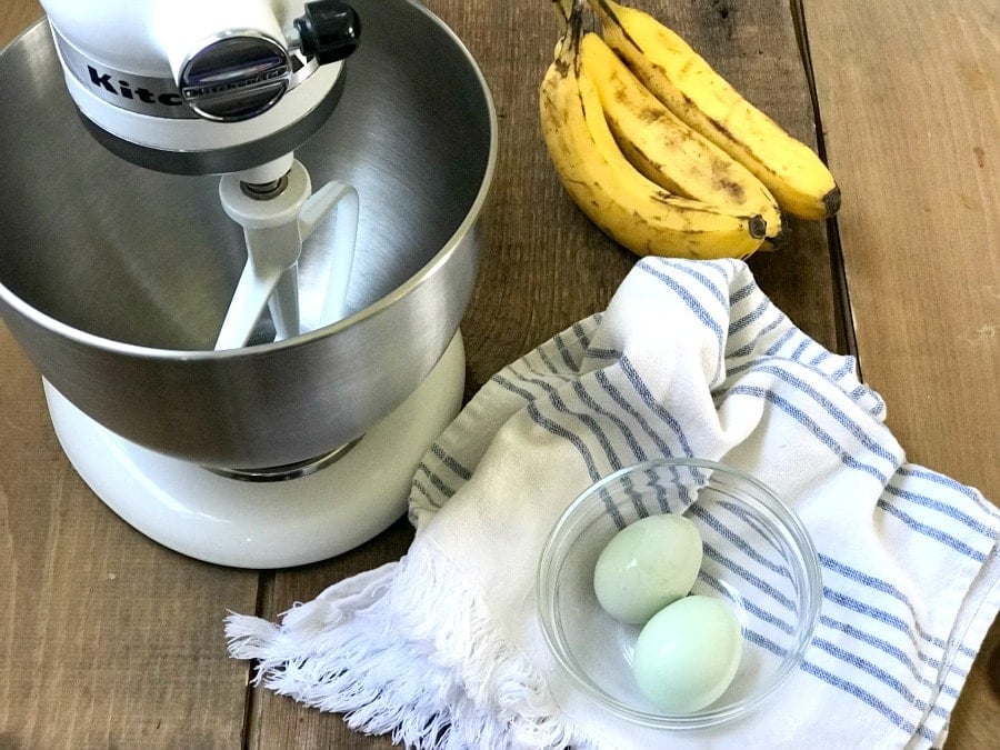 Overhead photo of two eggs in a glass bowl on a blue and white striped kitchen towel, three bananas, and a stand mixer on a wooden surface.