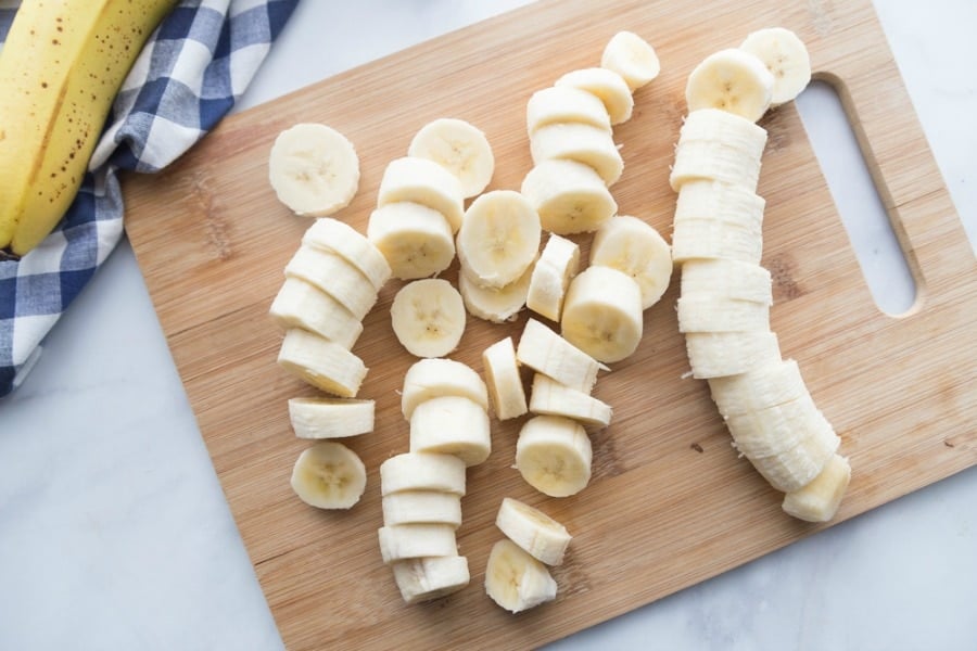 Sliced bananas on a wooden cutting board on a white marble surface. In the corner you can see part of a banana on a blue and white gingham towel.