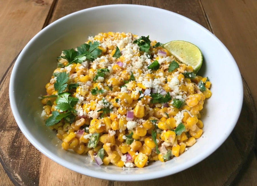 A white bowl filled with Mexican street corn salad topped with cotija cheese and cilantro with a lime wedge on the side