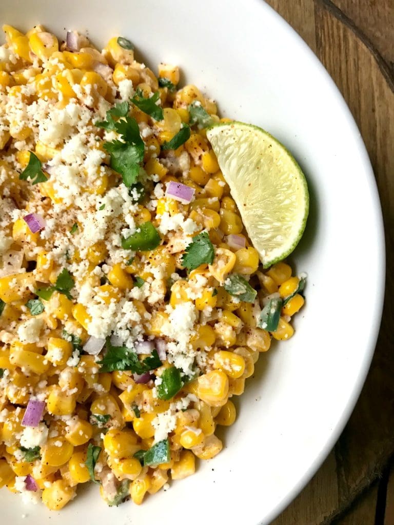 A white bowl filled with Mexican street corn salad topped with cotija cheese and cilantro with a lime wedge on the side