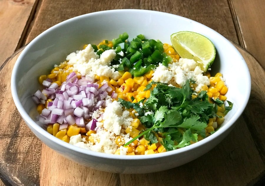 A white bowl containing corn, red onion, pepper, cotija cheese, lime, and cilantro- the ingredients for mexican street corn salad- on a wooden surface