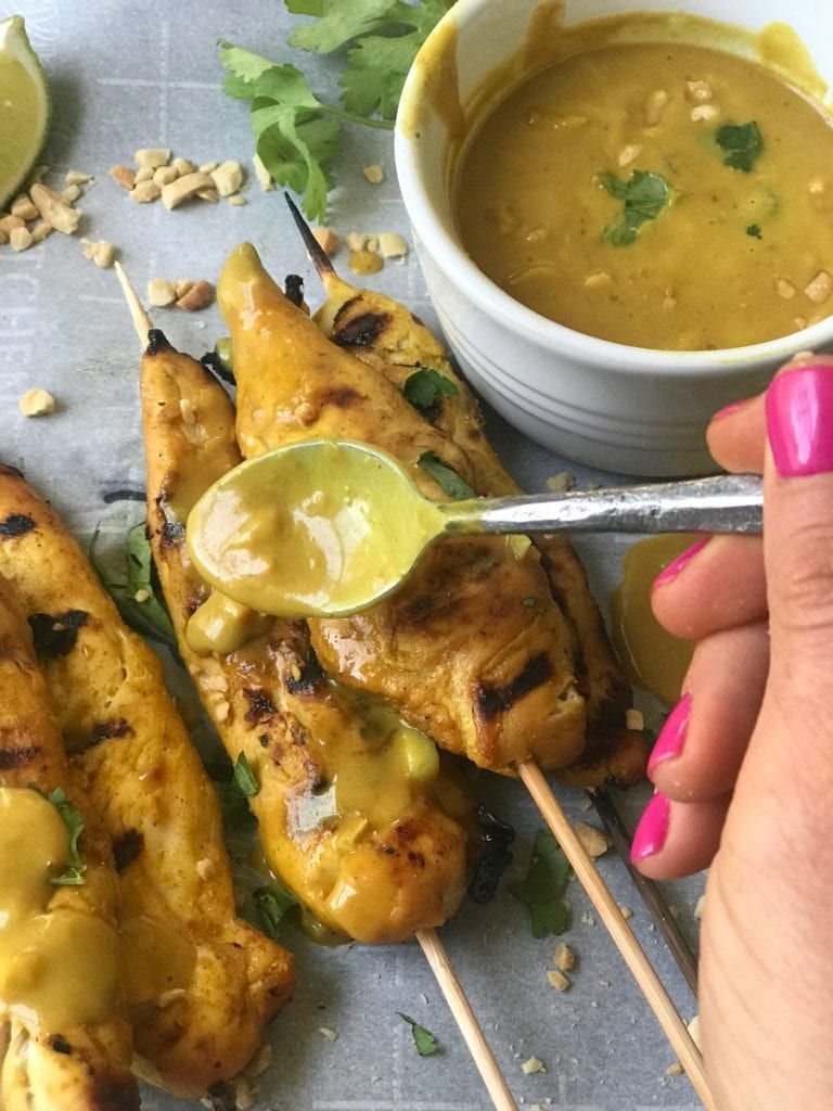 Chicken satay skewers drizzled with creamy peanut sauce dripping off spoon. A dish of the sauce is off to the side of the parchment lined tray.