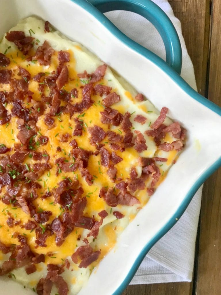 A square baking dish with mashed cauliflower topped with bacon, cheese, and chives