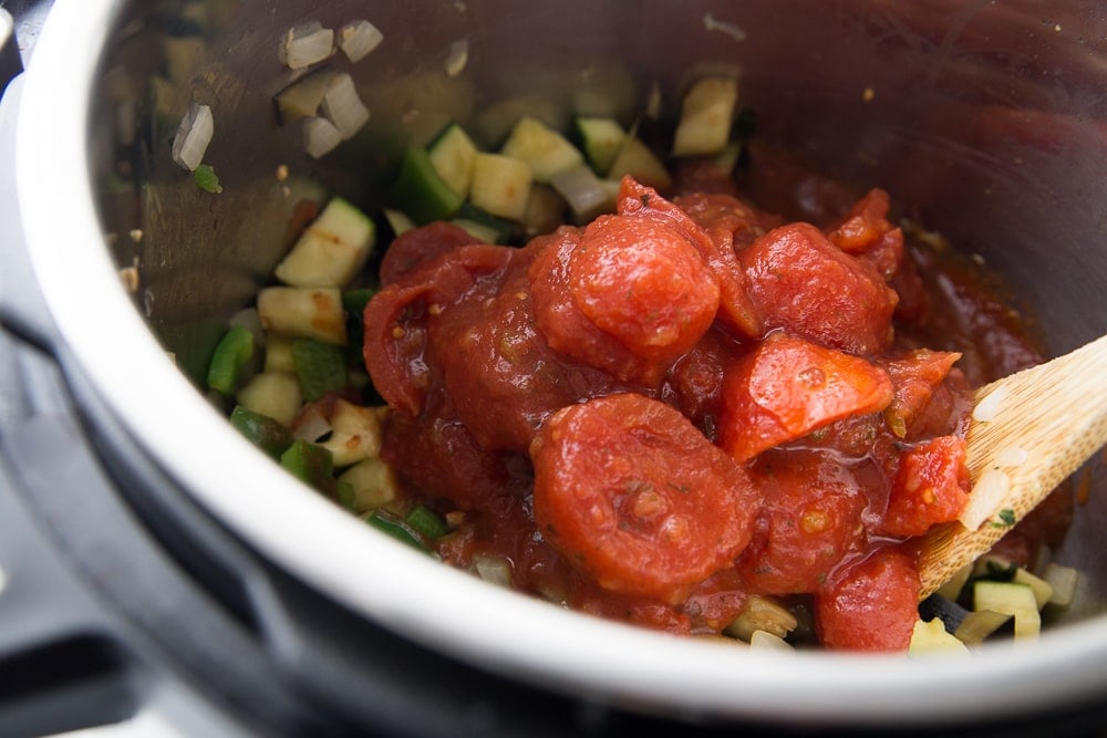 An instant pot full of veggies with a pile of canned tomatoes just added - wooden spoon resting on the side, ready to stir