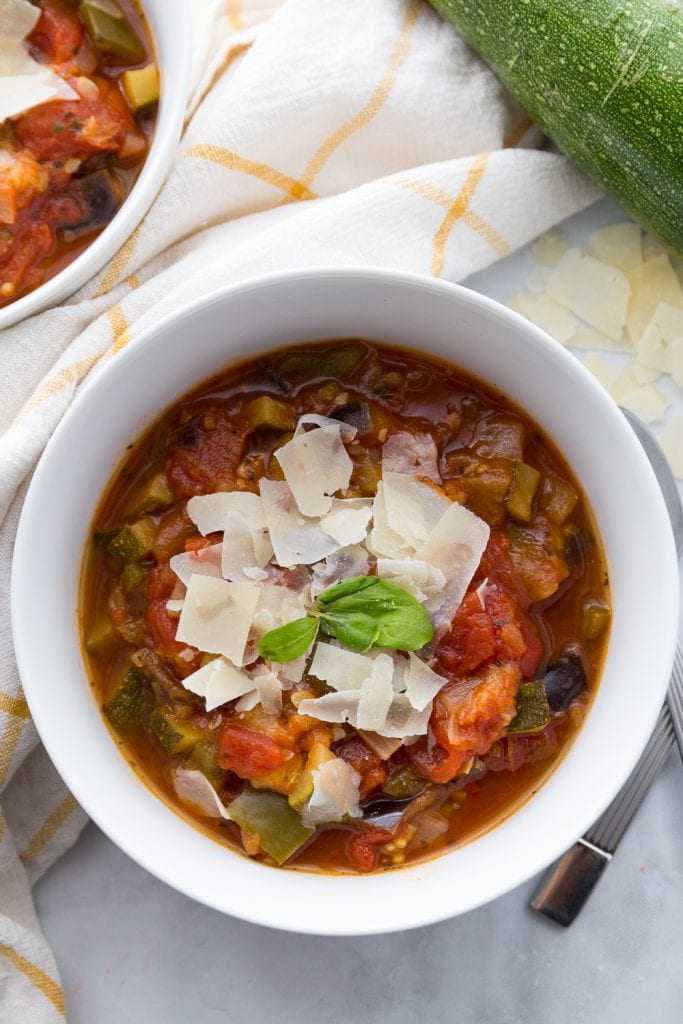 Two white bowls full of veggie packed ratatouille topped with Parmesan cheese and basil with a white and yellow plaid towel in the background