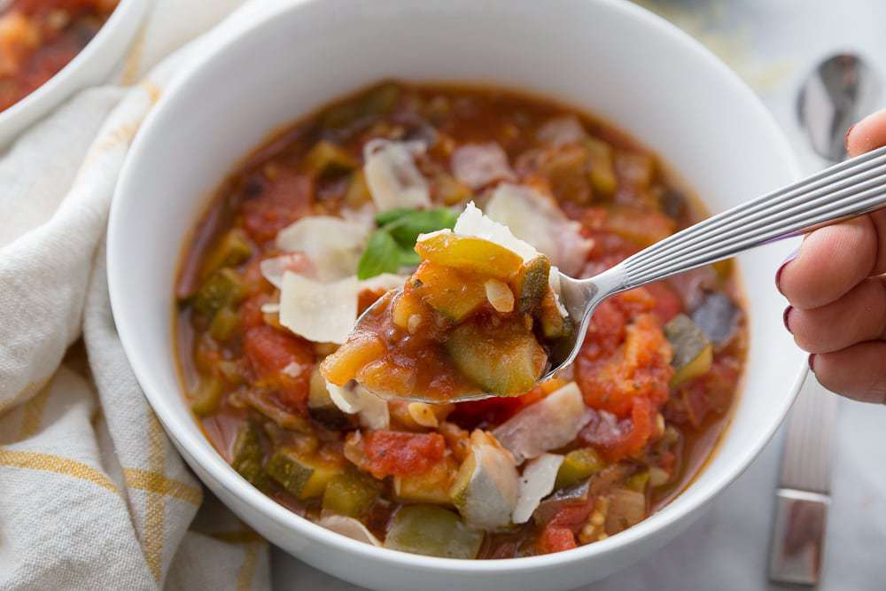 A spoonful of veggies is poised for eating- a portion of ratatouille topped with Parmesan is in a white bowl in the background.