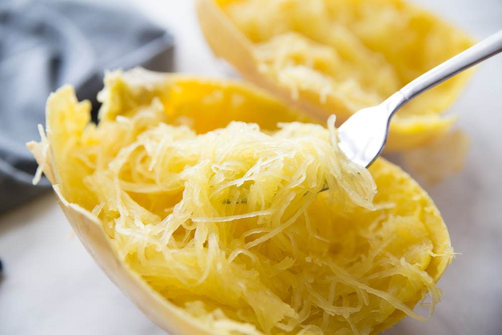 Cooked Spaghetti Squash Cut in Half with a fork showing the yellow strands of squash spaghetti on a while background 