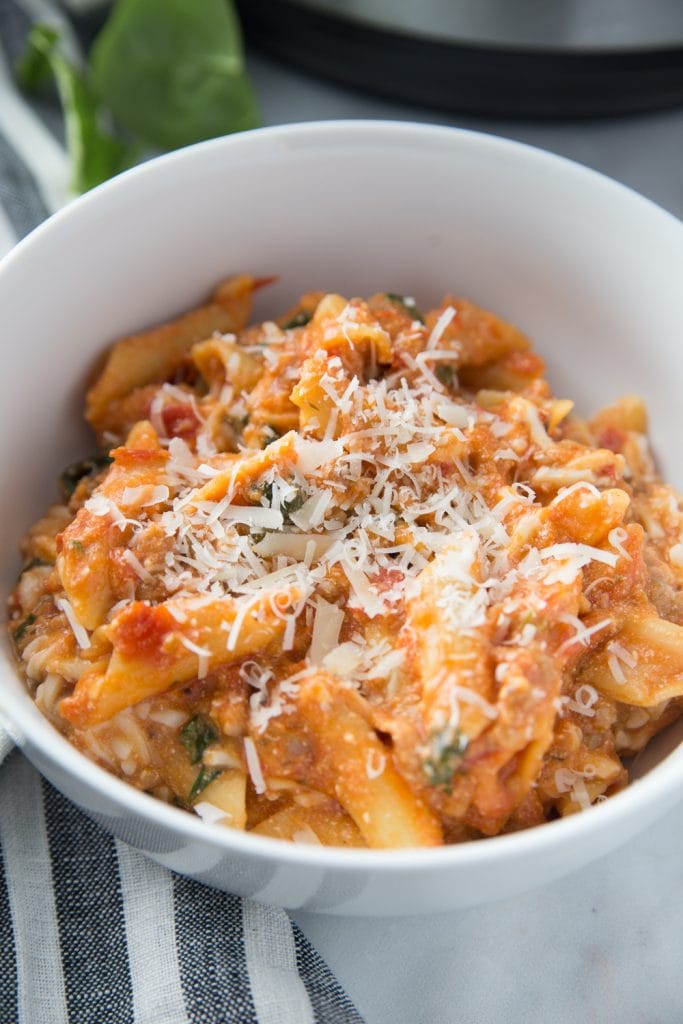 Close up of white bowl with baked ziti topped with cheese sitting on a blue and white striped kitchen towel. 