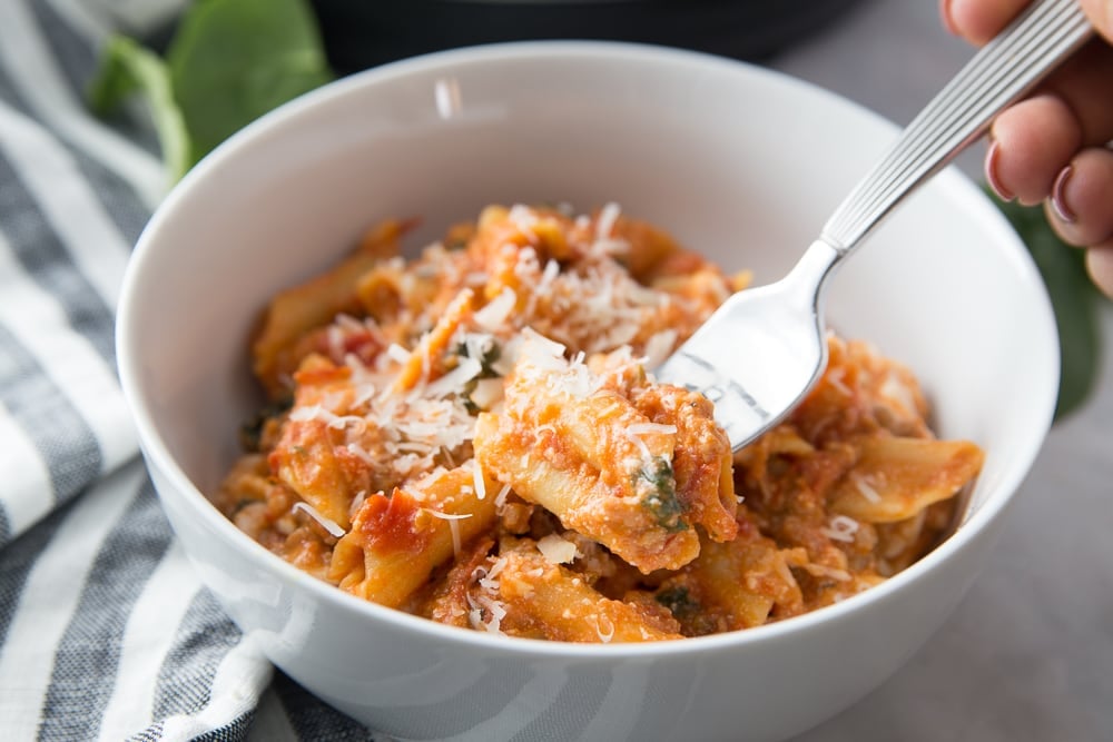 White bowl of baked ziti with someone's hand ready to take a bite with a silver fork 