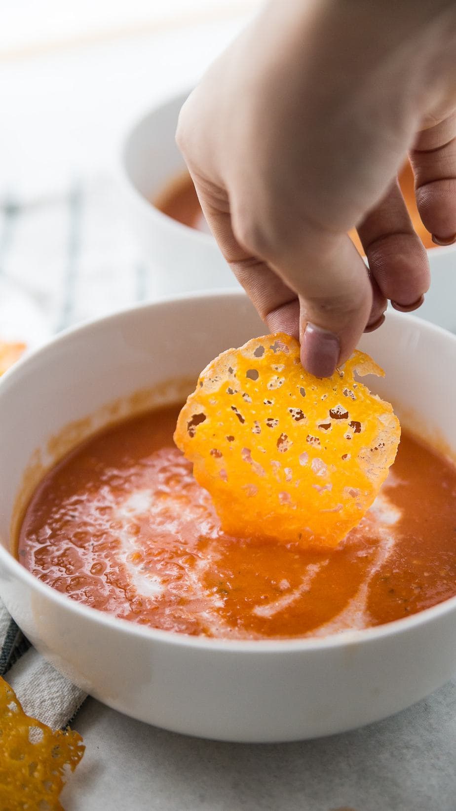 A woman's hand dipping a homemade crispy cheese chip into a white bowl of creamy tomato soup