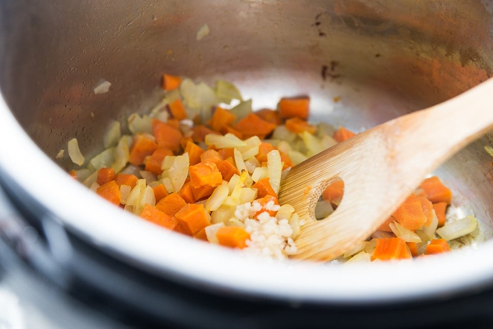 Instant Pot with carrots, onions, and garlic sauting. The Veggies are being stirred with a wooden spoon. 
