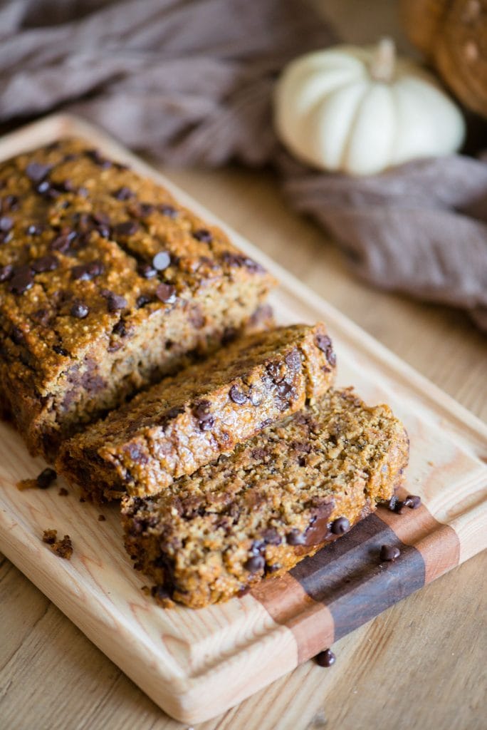 skiveskåret græskar havregrynbrød med chokoladechips på et træskærebræt. Det lyse træbord er prydet med hvide og orange græskar og et lysebrunt stykke stof.