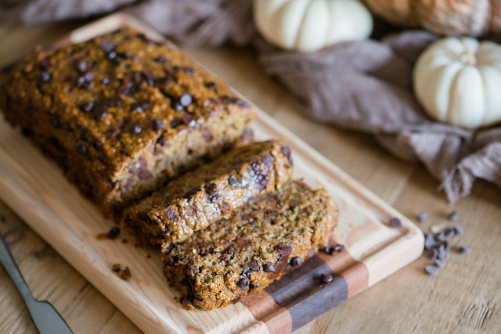Sliced Pumpkin Oatmeal bread with chocolate chips laying on a wooden cutting board. The light colored wood table is adorned with white and orange pumpkins and a light brown piece of fabric. A small knife sits beside the board. 