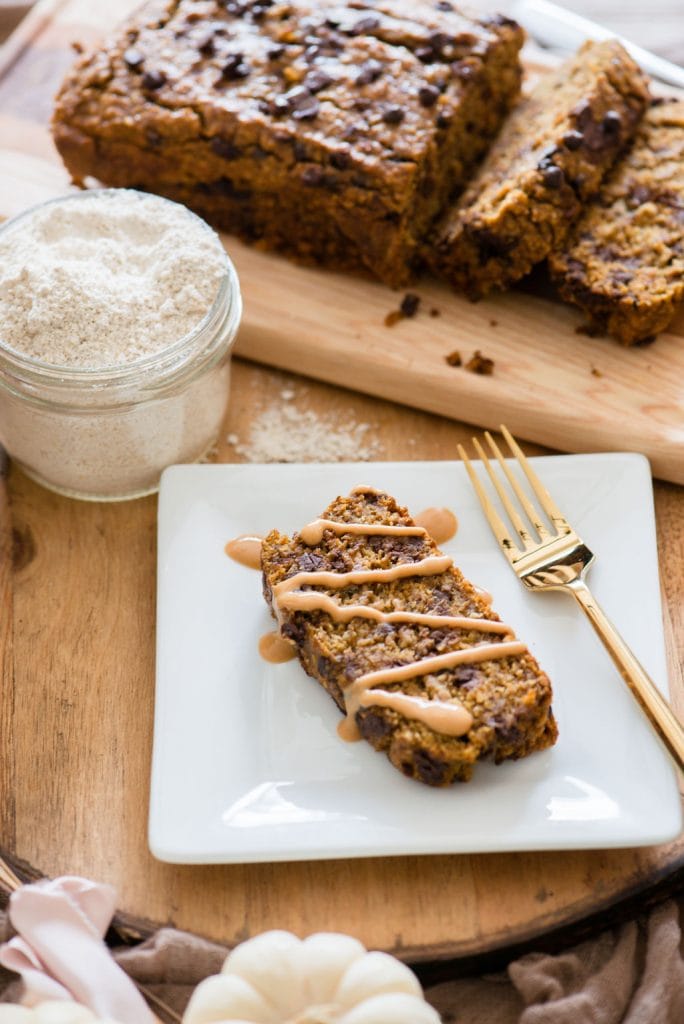 A loaf of Pumpkin Banana Oatmeal Bread sits on a wooden tray. Next to the bread is a small mason jar of oat flour and a plate with a slice of the bread with peanut butter drizzled on it. 
