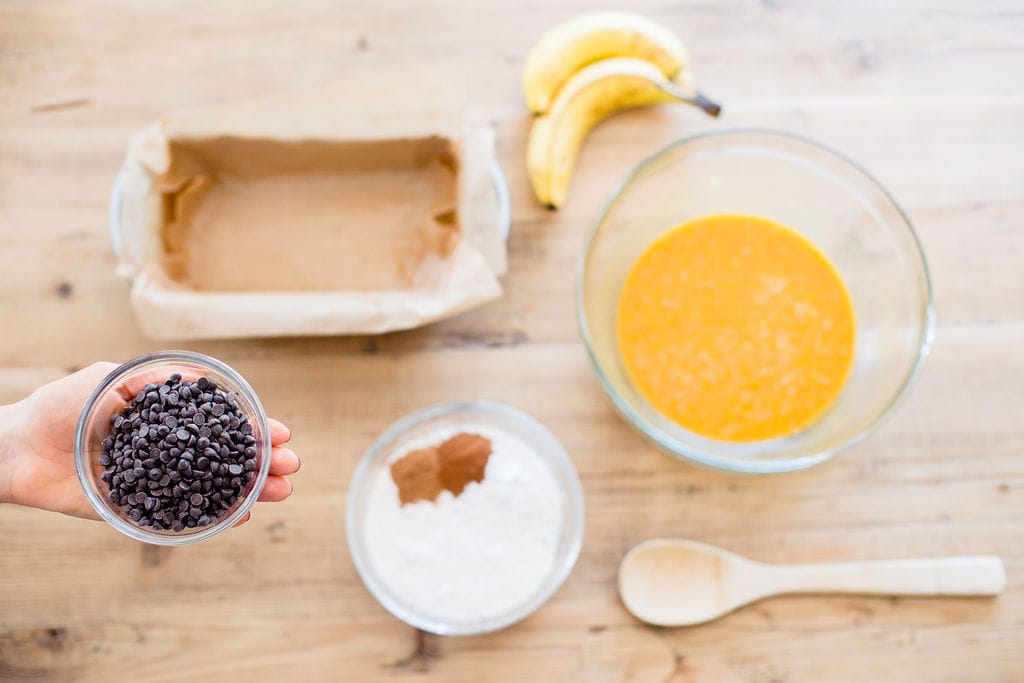 Una capa plana de los ingredientes para hacer Pan de Plátano con Harina de Avena y calabaza sobre una mesa de madera clara.