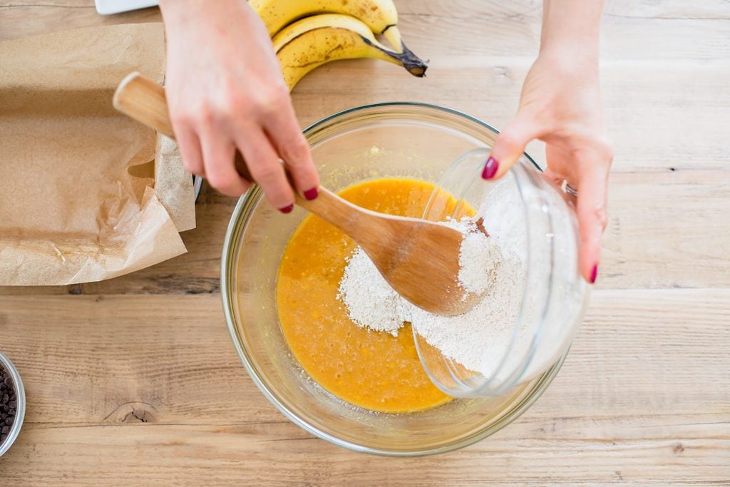 Mixing dry ingredients for Banana Pumpkin Oatmeal Bread into the wet ingredients. One hand is holding the small glass bowl and the other hand is stirring with a wooden spoon. Bananas are laying on the table, as well as a bowl of chocolate chips and a loaf pan with parchment paper. 