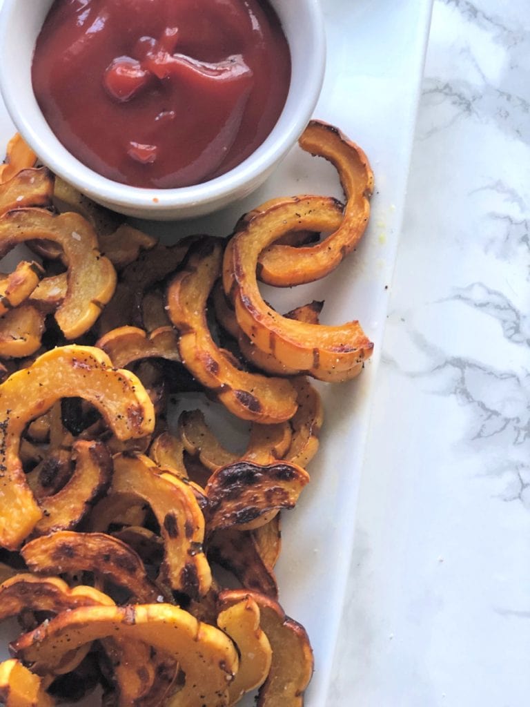 Roasted delicata squash fries on a white plate sitting on a marble table with a side of healthy ketchup. 