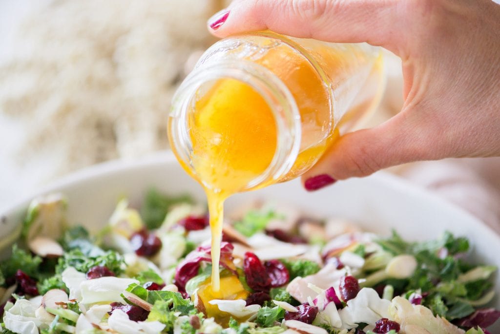 Close up of a woman's hand pouring maple cider vinaigrette out of a glass jar onto a bed of festive Holiday Salad.