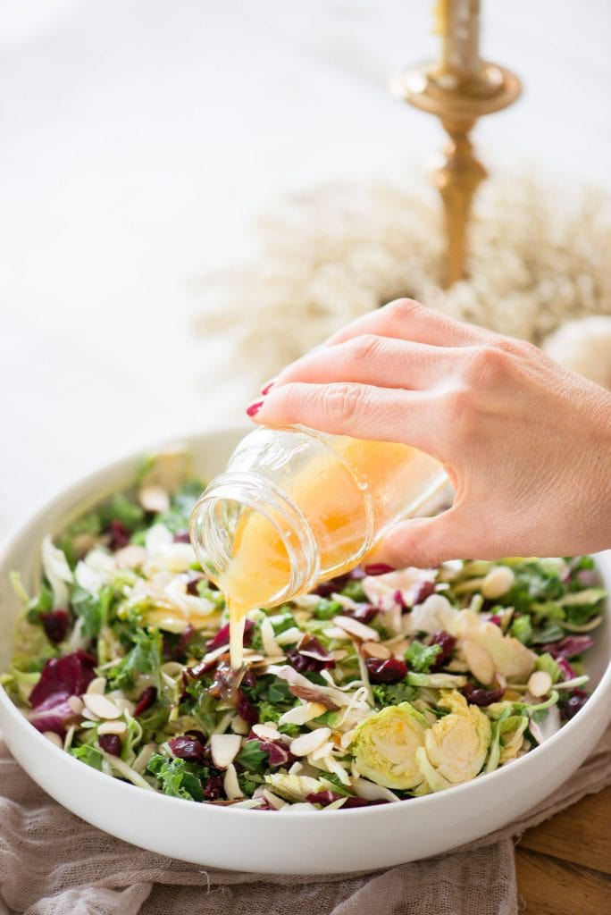 A woman's hand pouring maple cider vinaigrette out of a glass jar onto a bed of festive Holiday Salad. The salad made of purple cabbage, shaved Brussels sprouts, broccoli slaw, kale, and topped with dried cranberries and sliced almonds, is in a large white serving bowl. In the background is gold and cream fall decor.