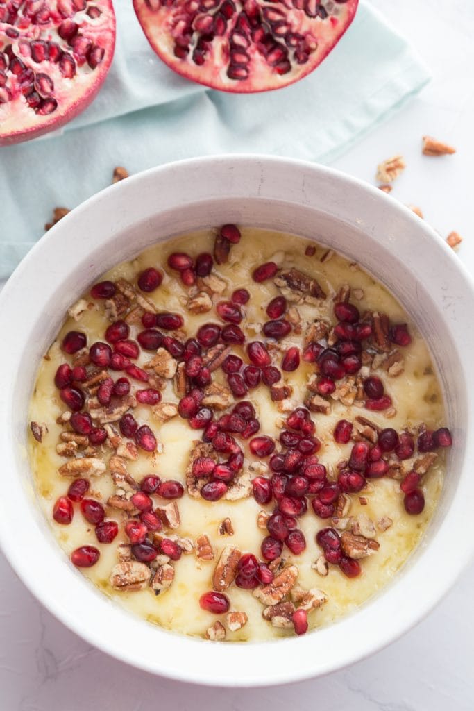 Overhead photo of a white ceramic bowl of melted brie with pomegranate seeds, honey and pecans. Two pomegranate halves sit nearby