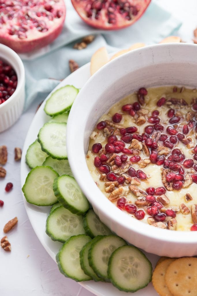 A white ceramic bowl of melted brie with pomegranate seeds, honey and pecans. Crackers and Cucumber dippers are sitting nearby. 