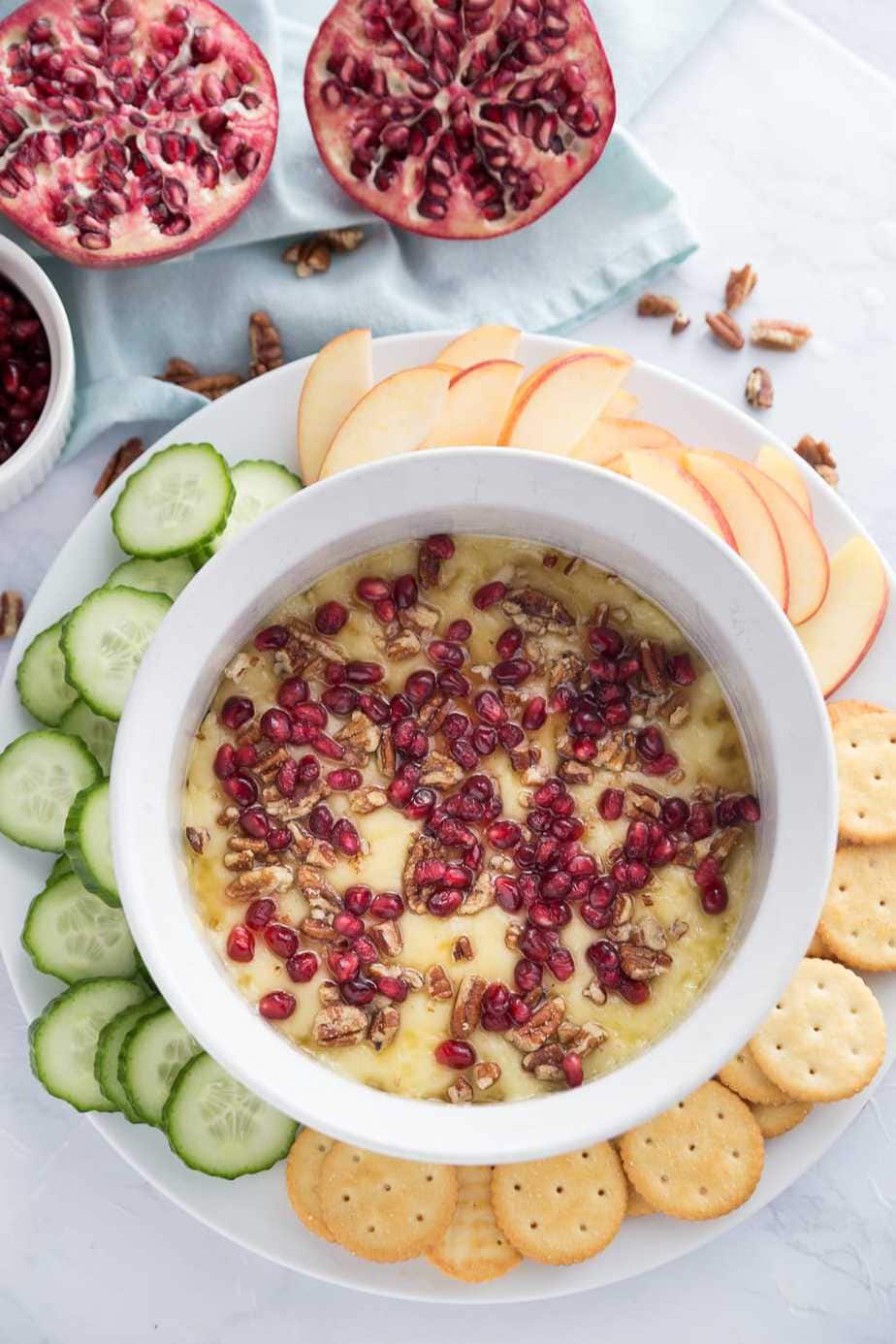 Overhead photo of baked brie appetizer topped with nuts and pomegranate seeds, in a white casserole dish. The casserole dish is sitting on a round white plate and cucumber slices, crackers, and apple slices surround the casserole dish. In the background is a pomegranate cut in half on a white napkin.
