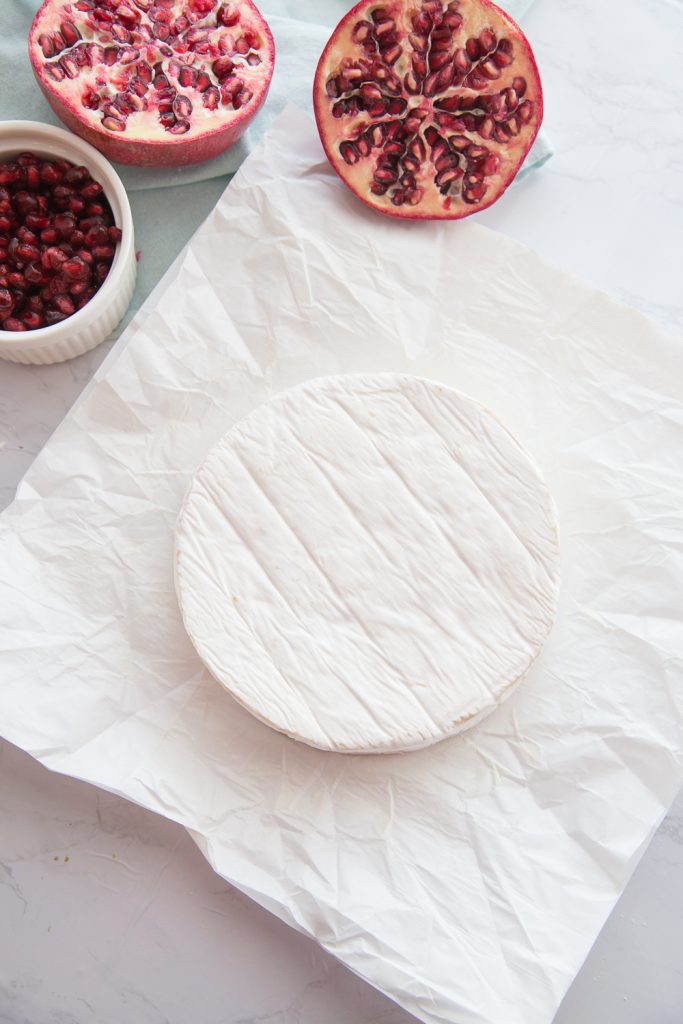 A brie wheel is unwrapped from it's white paper with a halved pomegranate on the side and a white bowl of pomegranate arils..