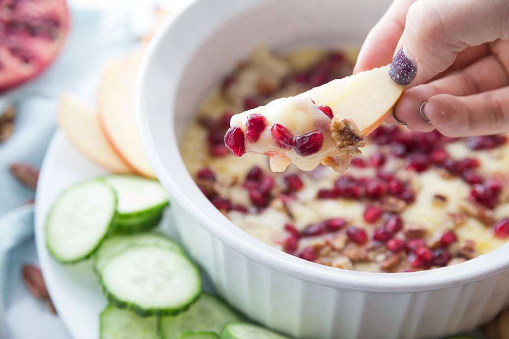 A white ceramic bowl of melted brie with pomegranate seeds, honey and pecans. A hand is holding an apple slice covered in brie. 