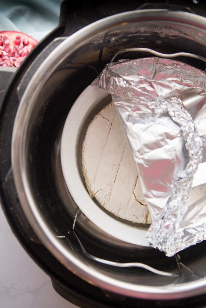 An Instant Pot with a white ceramic dish inside with a wheel of brie half covered in aluminum foil