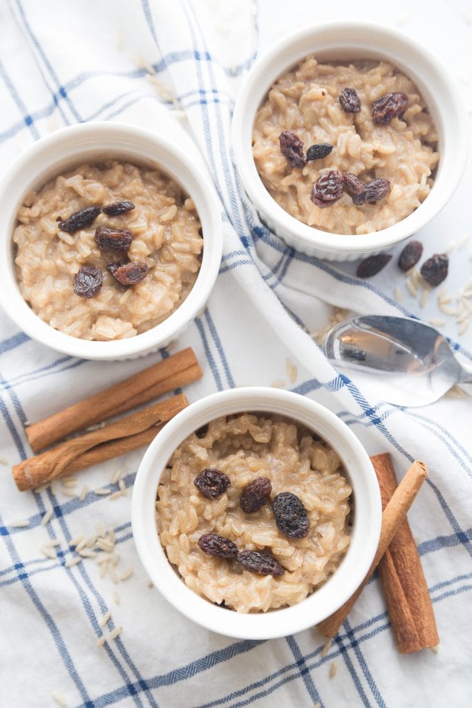 Three ramekins with healthy brown rice pudding, sitting on a blue and white cloth napkin. The rice pudding is topped with raisins and cinnamon, and cinnamon sticks sit nearby. 