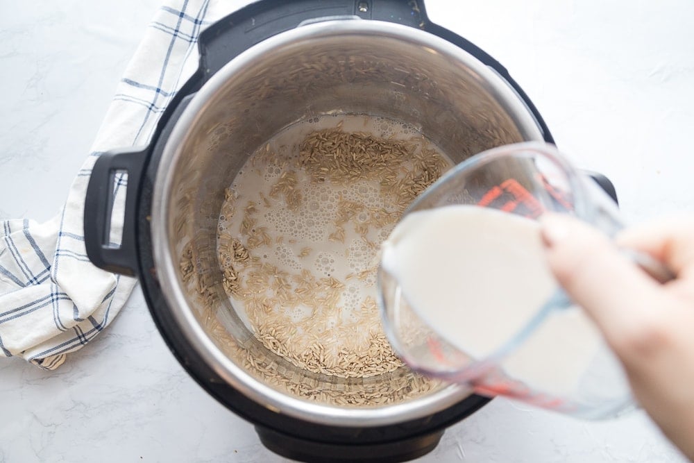 Overhead photo of uncooked rice in an instant pot with milk being poured into the pot from a clear measuring cup. On the side is a blue and white plaid kitchen towel on a white marble surface.