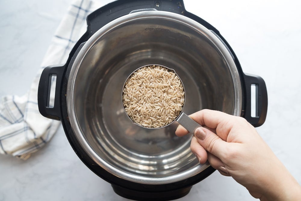 An Instant Pot sitting on a white marble counter. A woman's hand is holding a cup of brown rice over the pot. 