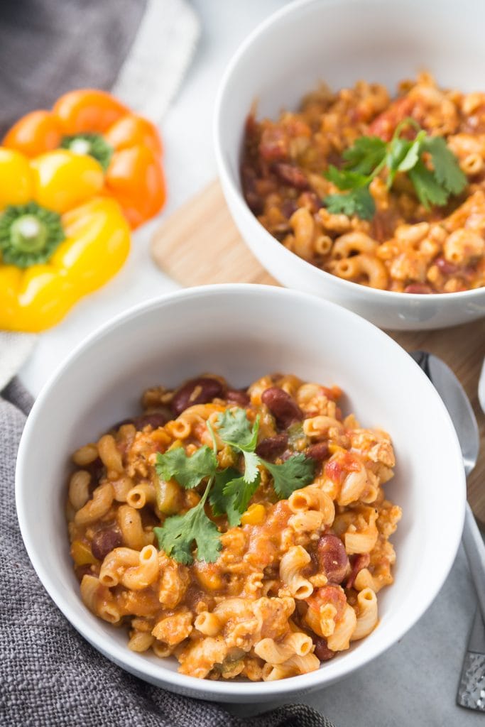 Two bowls of healthy chili mac sit side by side in white bowls topped with fresh cilantro. Bell peppers are laying next to the bowls on a wooden cutting board. 