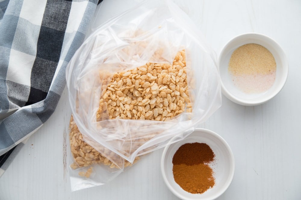 A large ziplock bag of brown rice cereal on a white background next to a checkered kitchen towel and two bowls of spices. 