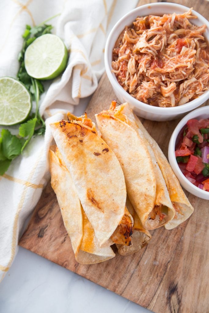 Cooked Air Fryer Chicken Taquitos next to a bowl of chicken and salsa with some lime nearby. 