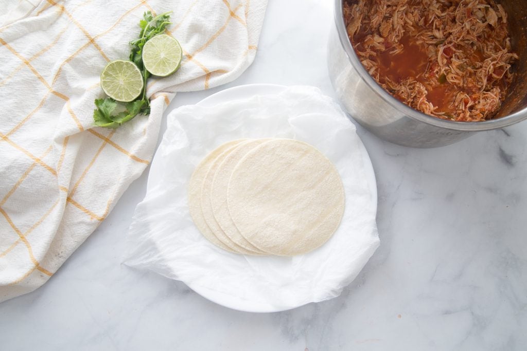 A plate of corn tortillas on a marble backdrop next to some limes and a bowl of salsa lime chicken 