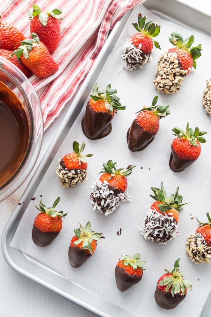 A tray of chocolate covered strawberries next to a bowl of melted chocolate. Some of the strawberries have shredded coconut and chopped peanuts