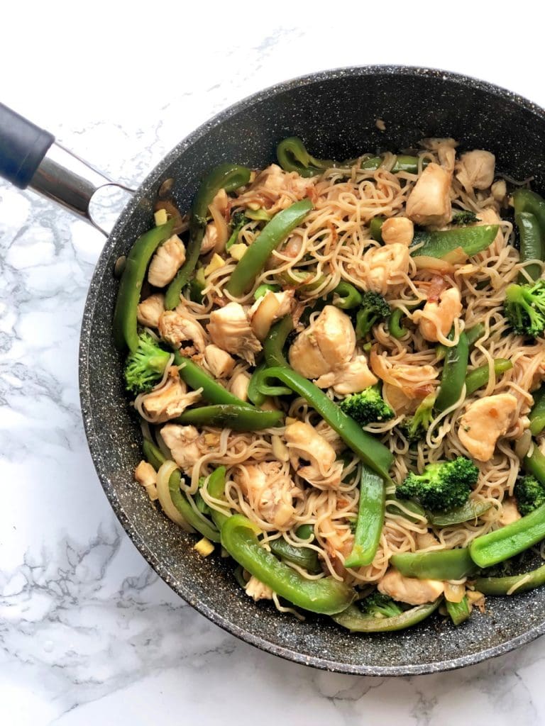 A skillet with Chicken Ramen Stir Fry with brown rice ramen, bell peppers, onions, and broccoli. The skillet sits on a marble background. 