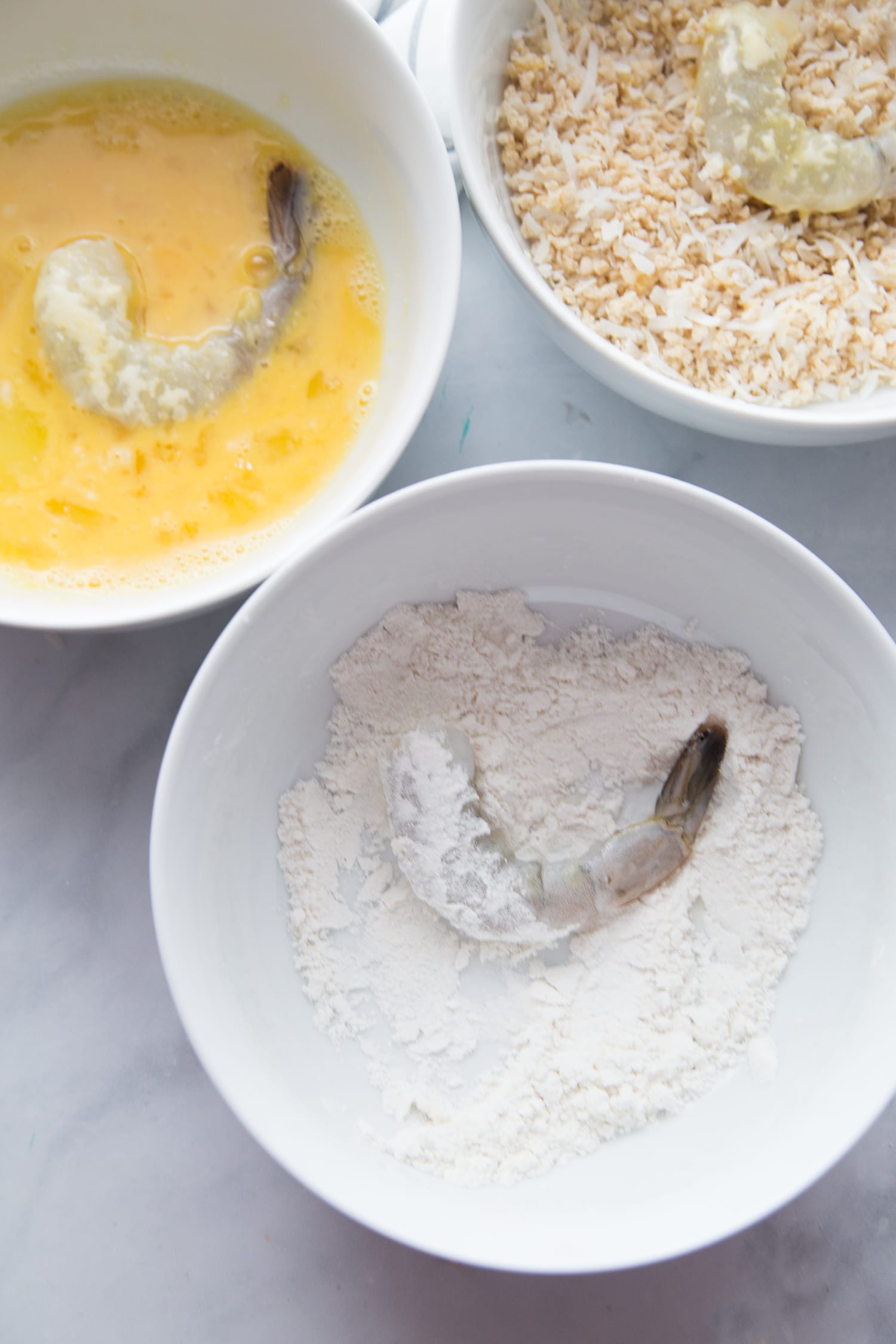 Three white bowls on a marble backdrop - one has egg with a shrimp, one has gluten free flour with shrimp, and one has panko breadcrumb with shrimp. 