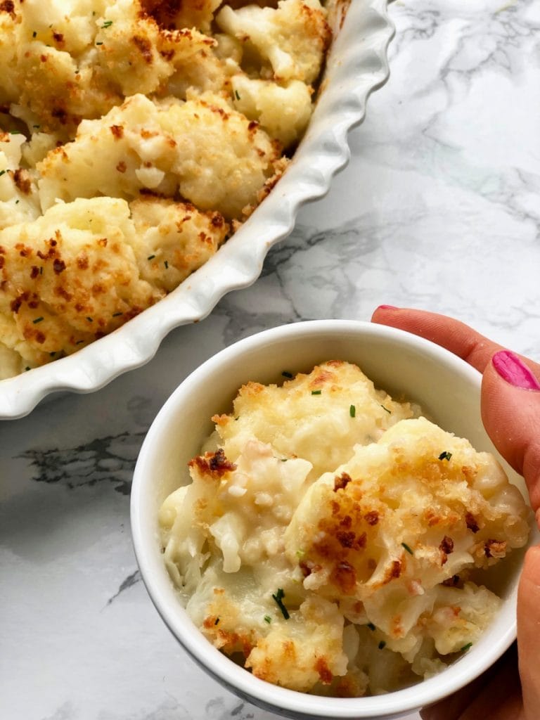 A woman's hand is holding a small bowl of Cauliflower Au Gratin next to a larger casserole dish of the same recipe. 