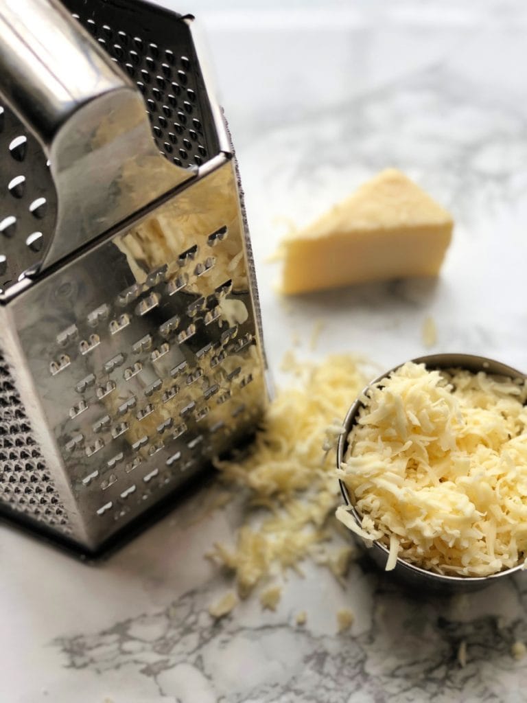 View from above of a box cheese grater next to a measuring cup full of shredded cheese and a wedge of cheese on a marble surface. This shredded cheese is ready for the freezer.