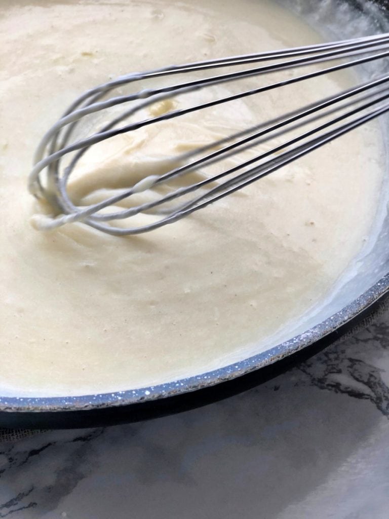 A white creamy cheese sauce is being whisked in a skillet using a wire whisk 