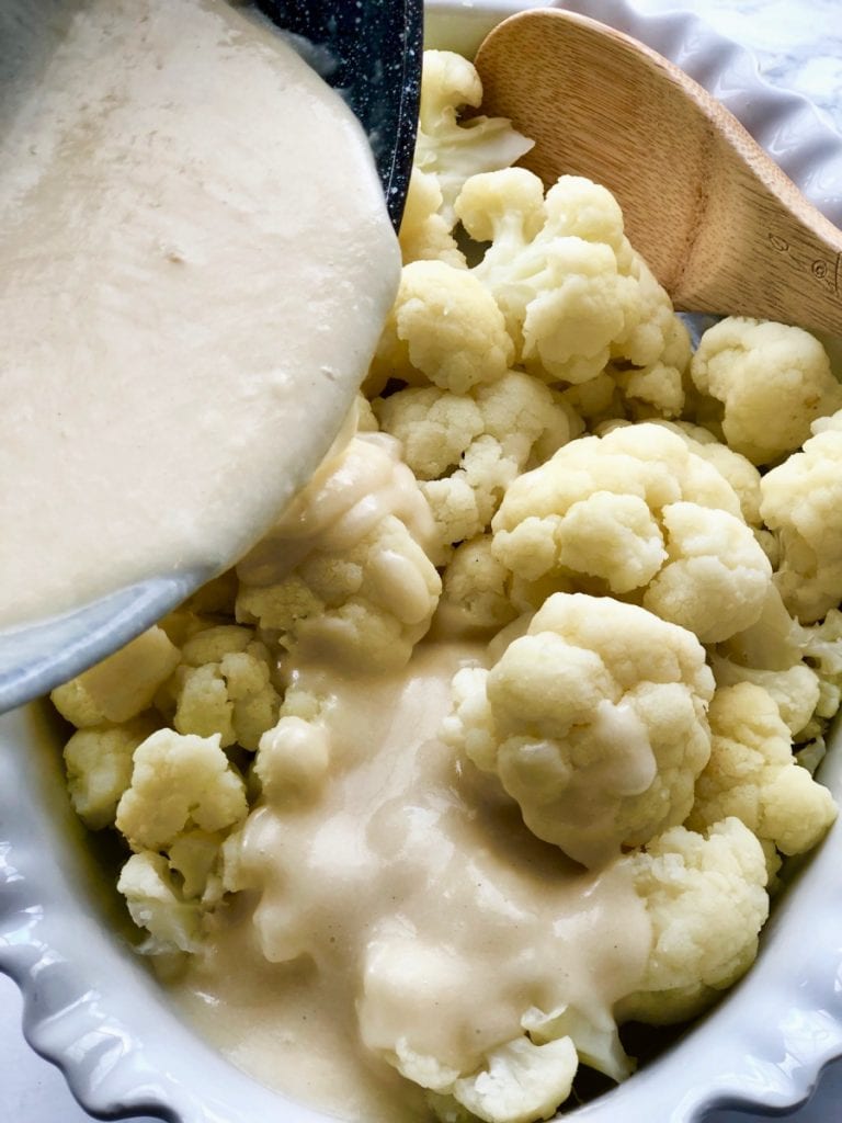 A white creamy cheese sauce is being poured over cauliflower florets in a white oval casserole dish